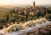 Dove Cameron and Damiano David Wedding: Couple Marry in Montalcino Hilltop Tuscan town at golden hour above vineyards, with a wedding table of white hydrangeas and candles on a stone terrace in the foreground.