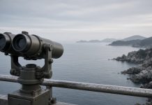 North Korea Fires Ballistic Missile After Seoul Demands Apology Over DMZ Mine Blast Mounted observation binoculars on a metal railing overlooking a calm grey sea and a rocky coastline under an overcast morning sky