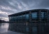 Manchester City Lodge Appeal Against Premier League Guilty Verdict: What Happens Next A modern football stadium with a curved glass facade and spiral access towers at dusk in the rain, with an empty wet plaza in front