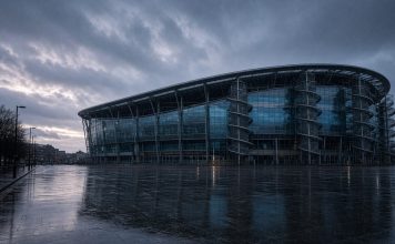 Manchester City Lodge Appeal Against Premier League Guilty Verdict: What Happens Next A modern football stadium with a curved glass facade and spiral access towers at dusk in the rain, with an empty wet plaza in front